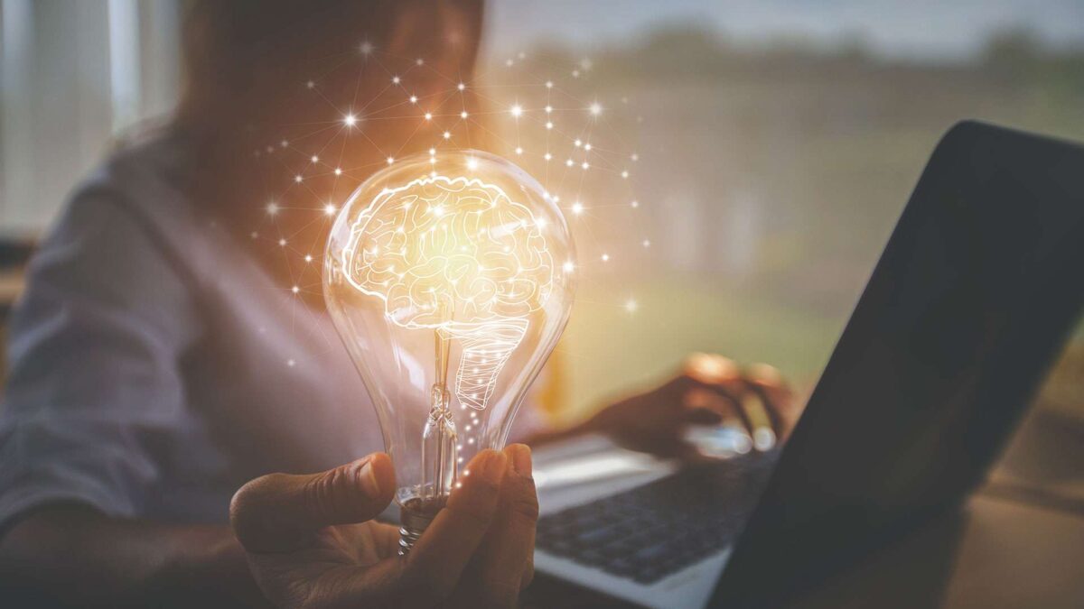 A person holds a glowing lightbulb with a brain design inside, symbolizing ideas or innovation, while working on a laptop. The background is softly lit, hinting at a thoughtful or creative environment.