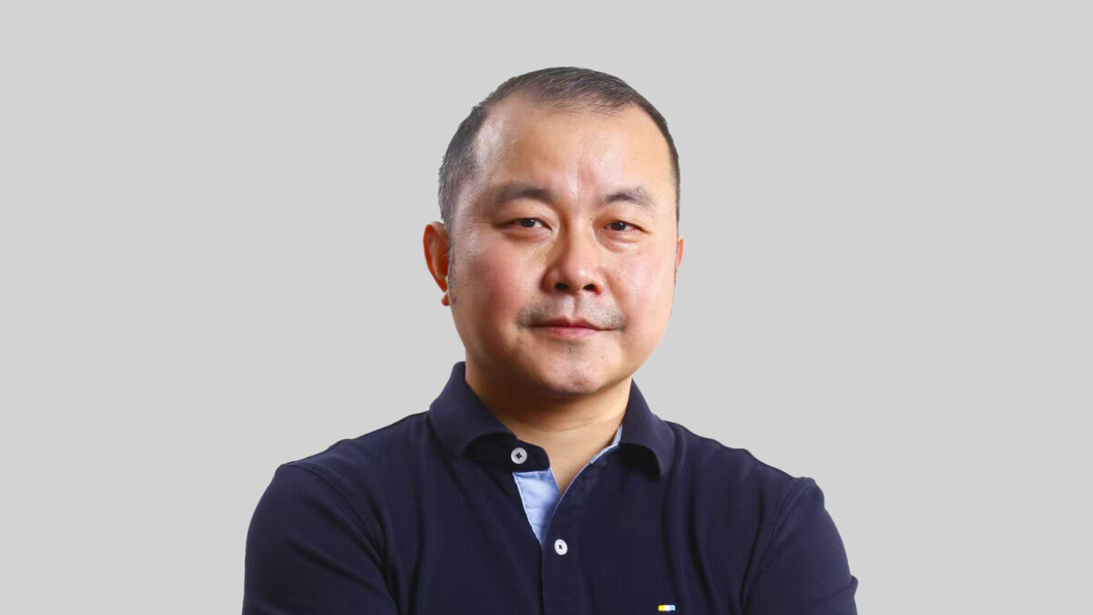 A person with short hair and a neutral expression is standing against a plain background, wearing a dark polo shirt with crossed arms.