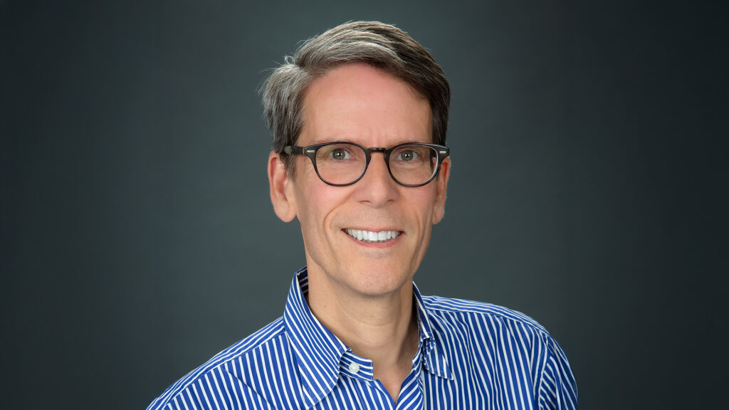 A person with short, gray hair and glasses smiles at the camera. They are wearing a blue and white striped shirt against a dark background.