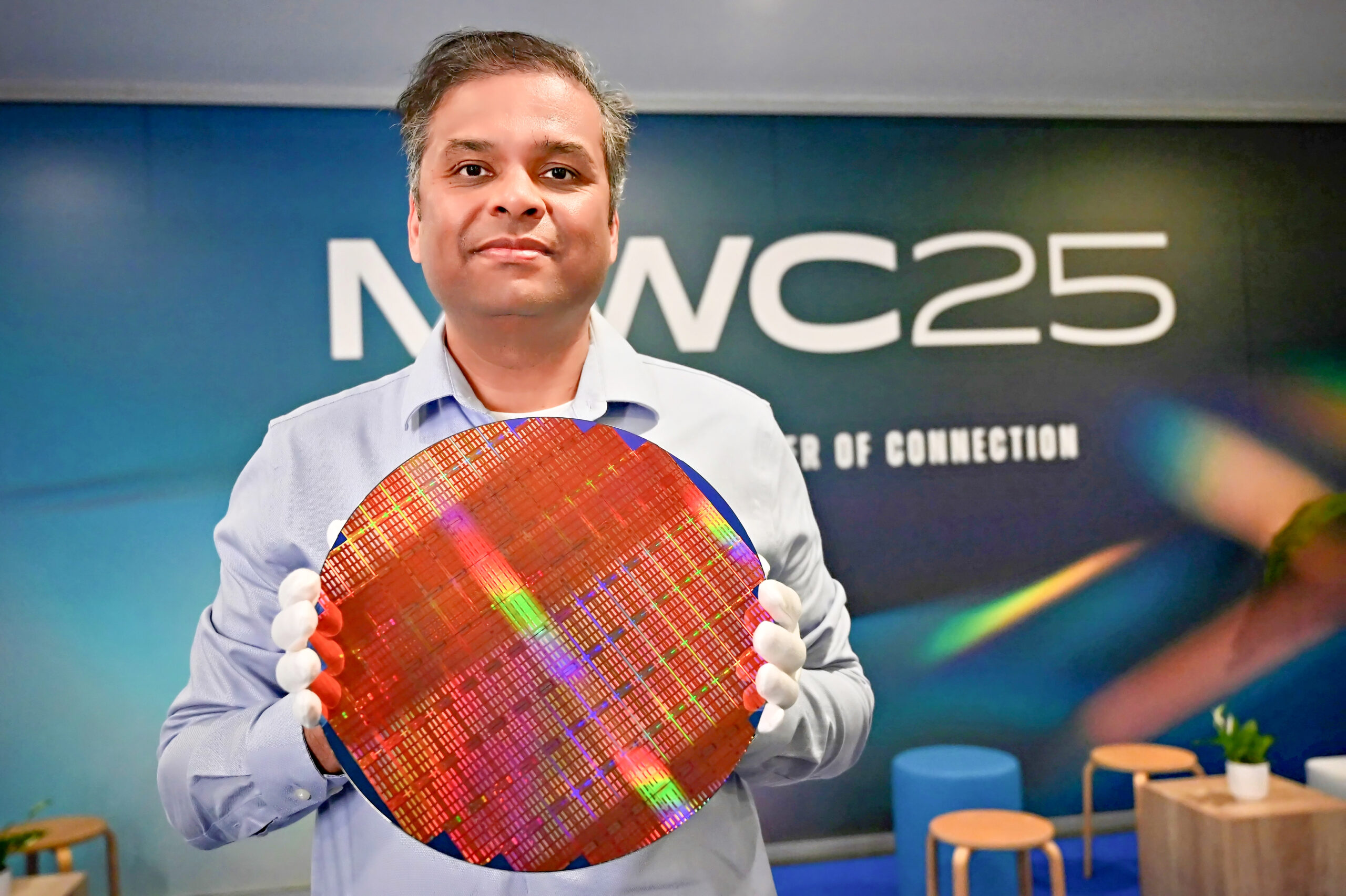 A person in a light blue shirt holds a large, circular silicon wafer with multicolored reflections. They stand in front of a blurred background with the text MWC25 The Power of Connection.