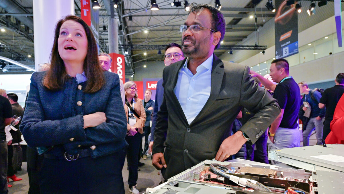 A woman and a man are standing together at a technology exhibition, surrounded by a crowd. They are observing open computer hardware on display. The event is busy, with various tech brand signs visible in the background.