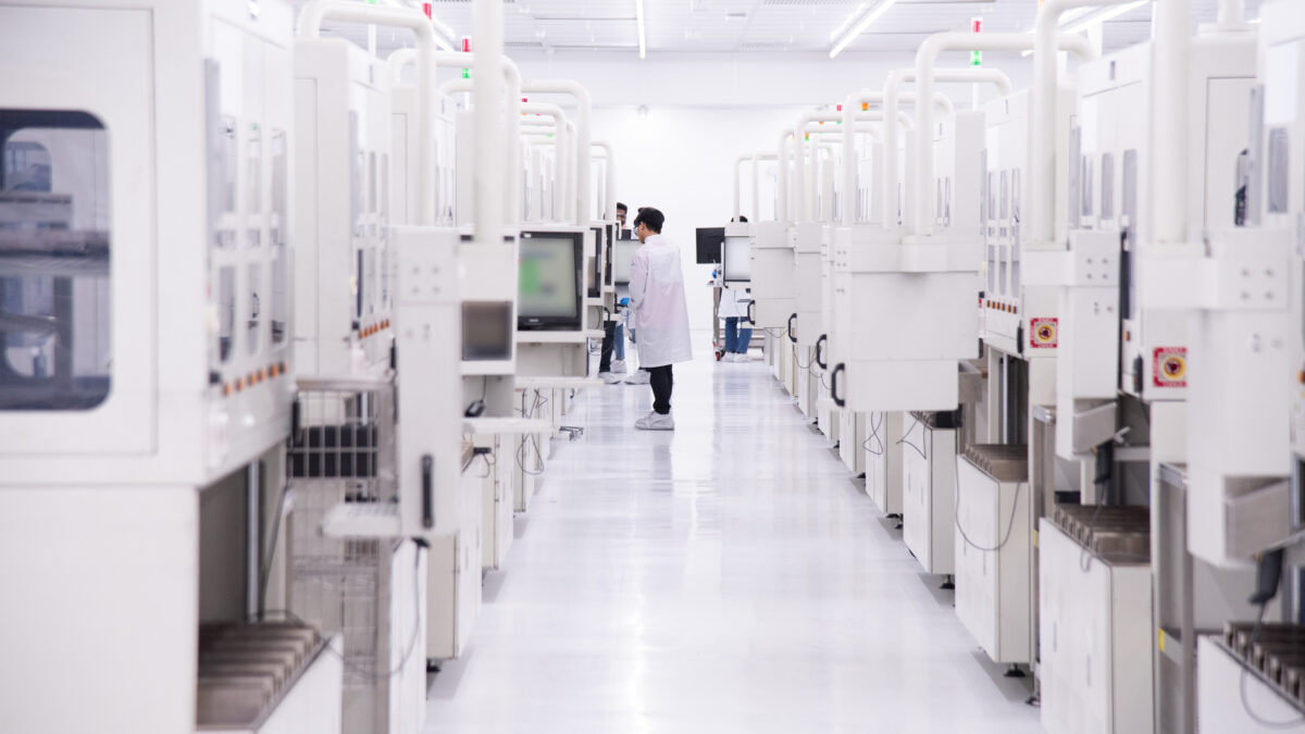 A person in a white lab coat stands in a bright, sterile laboratory with rows of tall, white equipment lining both sides of the aisle. They are looking at a screen, and the floor is shiny and reflective.