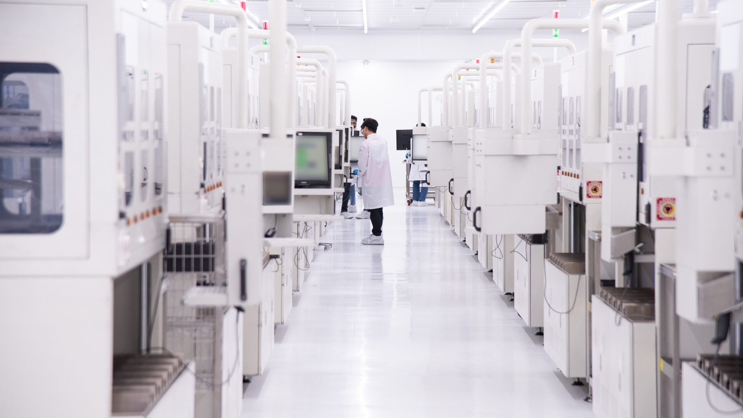 A person in a white lab coat stands in a bright, sterile laboratory with rows of tall, white equipment lining both sides of the aisle. They are looking at a screen, and the floor is shiny and reflective.