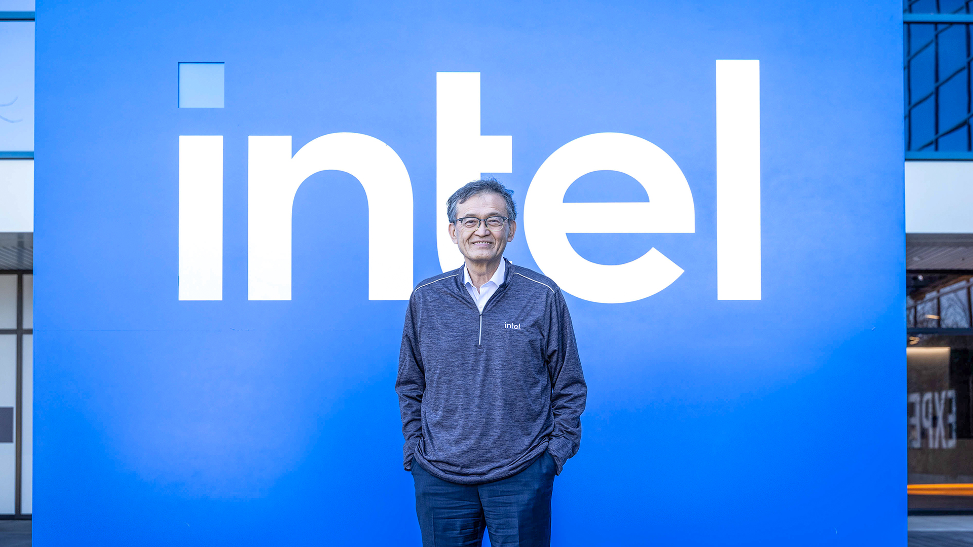 A person wearing a dark pullover stands in front of a large blue wall with the white Intel logo. They have their hands in their pockets and are smiling.