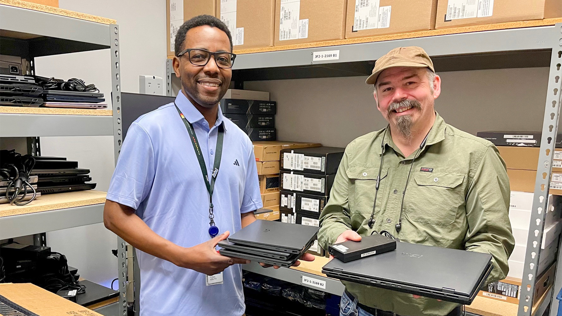 Two smiling men in a storage room hold laptops. One wears a light blue polo and glasses, the other a green shirt and hat. Shelves behind them are filled with boxes and more laptops.