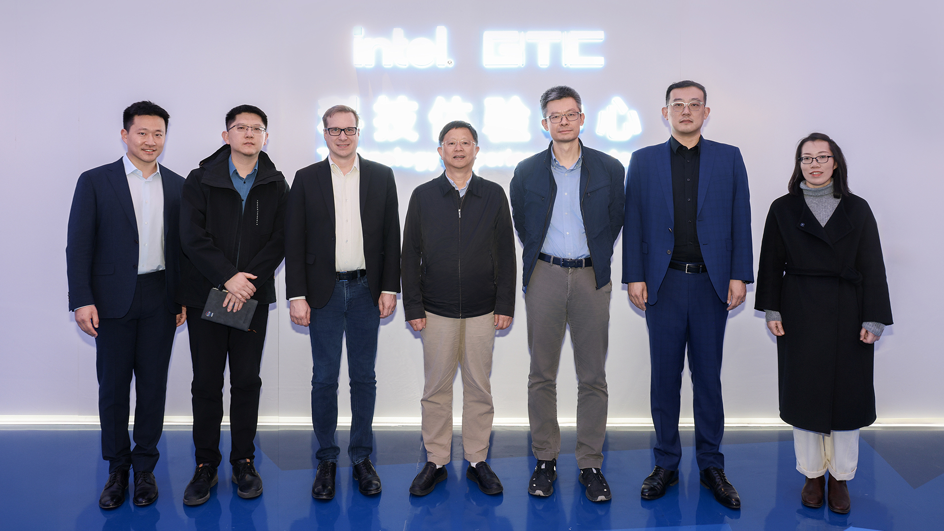 Seven people pose for a group photo against a backdrop with glowing text that reads intel GTC. They are dressed in business attire, standing in a line on a glossy floor.