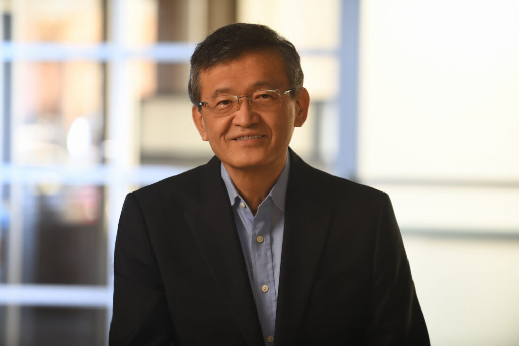 A man in a dark suit and light blue shirt smiles at the camera. He is wearing glasses and standing in front of a softly blurred background with a window grid pattern, giving the image a warm and professional atmosphere.