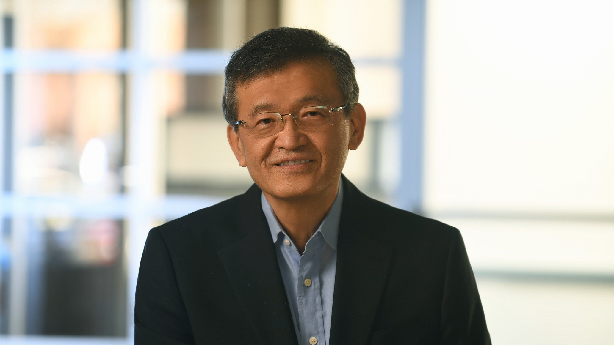 A person wearing glasses and a navy blazer over a light blue shirt smiles at the camera. They have short, dark hair and are in a softly lit indoor setting with blurred windows in the background.